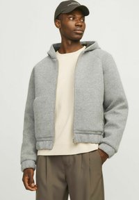 Grauer Reißverschluss-Hoodie aus weichem Fleece-Material mit einer Kängurutasche, gerippten Bündchen und Saum. Kombiniert mit einem cremefarbenen Crewneck-Shirt und braunen Hosen.