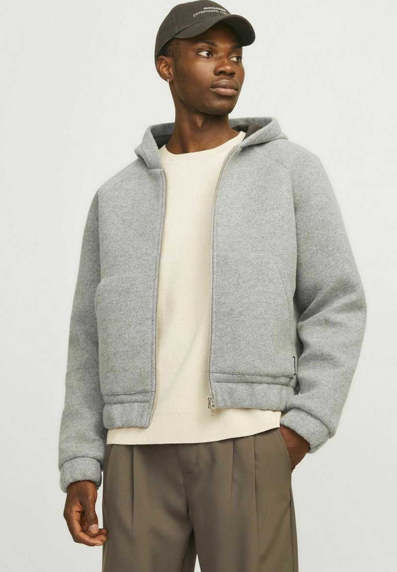 Grauer Reißverschluss-Hoodie aus weichem Fleece-Material mit einer Kängurutasche, gerippten Bündchen und Saum. Kombiniert mit einem cremefarbenen Crewneck-Shirt und braunen Hosen.