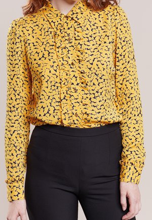 Kobieta ubrane w żółtą bluzkę z czarnym kwiatowym wzorem i falbankami, w połączeniu z czarnymi spodniami z wysokim stanem.