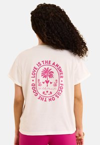 Camiseta de algodón blanca con un gran diseño rosa de una palmera, corazones y texto que dice "El amor es la respuesta" en la parte de atrás.