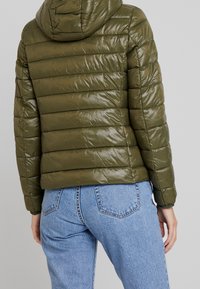 Olivgrön puffjacka med horisontella sömmar, huva och glänsande yta. Bärs över ljusblå högmidjade jeans.