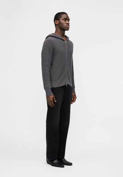 Άνδρας που φοράει γκρι πλεκτό φούτερ με φερμουάρ, μαύρ ο παντελόνι καμπάνα και μαύρα παπούτσια με μυτερή μύτη, στέκεται μπροστά σε λευκό φόντο.