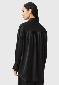 Blouse noire à manches longues en tissu lisse, dotée d'un col, de poignets boutonnés et d'un détail froncé au dos pour un ajustement décontracté.