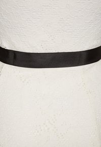 Biała koronkowa sukienka z teksturowanym wzorem oraz szerokim czarnym satynowym paskiem w talii. Skoncentrowano się na misternych detalach koronkowych.