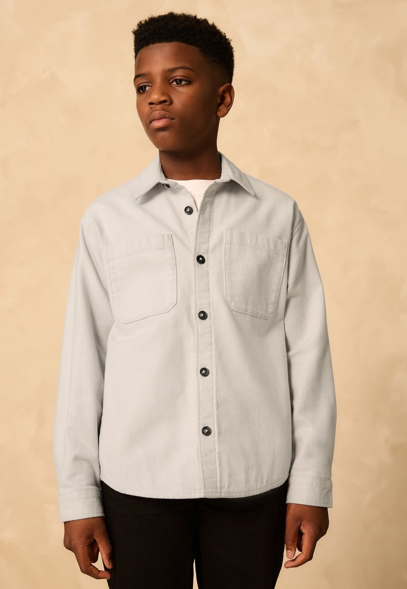 Chemise grise claire à boutons en tissu texturé, avec deux poches poitrine et des boutons noirs, associée à un pantalon foncé.
