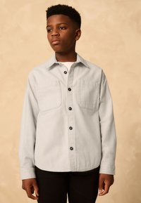 Light gray button-up shirt made of textured fabric, featuring two chest pockets and black buttons, paired with dark pants.
