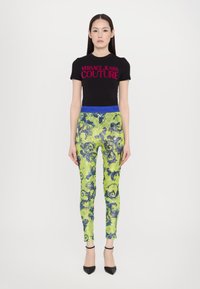Camiseta negra de manga corta con el logo en rosa "VERSACE JEANS COUTURE"; leggings verdes con lentejuelas y cintura azul, con un intrincado patrón floral azul.