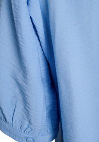 Camisa azul claro de botones con tela texturizada, puños fruncidos y un ajuste cómodo. El material parece suave y ligeramente arrugado.