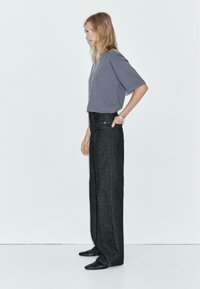 T-shirt grigio a maniche corte abbinato a jeans neri a vita alta e a gamba larga. Il modello sta in piedi di lato, indossando scarpe nere e un braccialetto.
