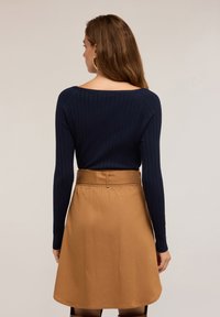 Frau mit langen braunen Haaren, die goldene Creolen trägt, ein navyblaues, geripptes Langarmoberteil und einen hoch taillierten kamelbraunen Rock, mit dem Rücken zur Kamera.