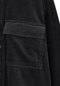 Black corduroy jacket with a textured surface, featuring a large front pocket and button fastenings. The design emphasizes the soft fabric's ribbing.