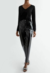 Suéter negro de cuello en V con tachuelas plateadas, combinado con pantalones de cuero negro brillantes. El calzado presenta un diseño de correa texturizada en plata.