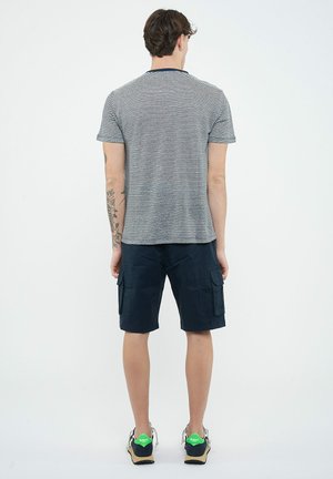 Giovane uomo con braccio sinistro tatuato che indossa una maglietta a righe bianche e nere, pantaloncini cargo blu navy e scarpe da ginnastica con talloni verdi, in piedi di spalle.