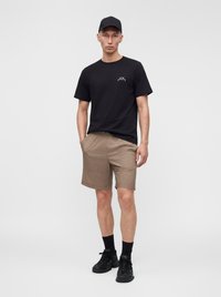 Jeune homme portant une casquette noire, un t-shirt noir, un short beige, des chaussettes noires et des baskets noires, debout les mains dans les poches.