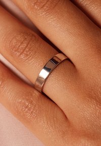 Polished silver band ring with a flat surface, gently rounding edges, worn on a hand with textured skin against a soft fabric background.