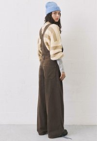 Braune Denim-Overalls mit lockerer Passform, ausgestattet mit aufgenähten Taschen und verstellbaren Trägern. Kombiniert mit einem gestreiften, flauschigen cremefarbenen Pullover und einer blauen Mütze.