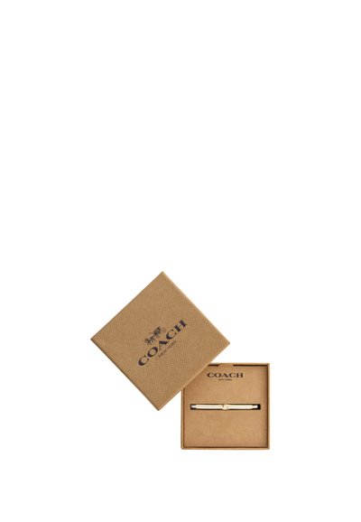 Une pince à cravate Coach New York dans un coffret de présentation beige ouvert, avec le couvercle assorti portant le logo de la marque posé à côté.