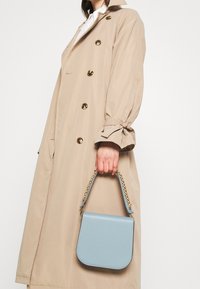 Square light blue handbag with a gold chain strap, made of smooth material. Held by a person wearing a beige trench coat with gold buttons.