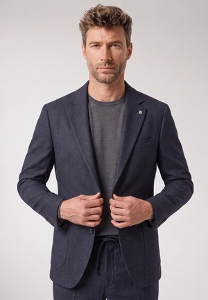 Man draagt een donkerblauw colbert over een grijs overhemd en stelt het colbert bij, staand tegen een effen lichte achtergrond.