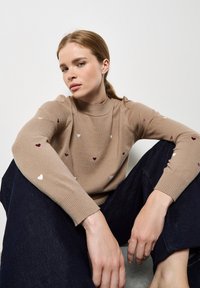 Beige trui met lange mouwen, versierd met kleurrijke hartborduursels; geribbelde hals en boorden, gecombineerd met donkere wijde jeans.