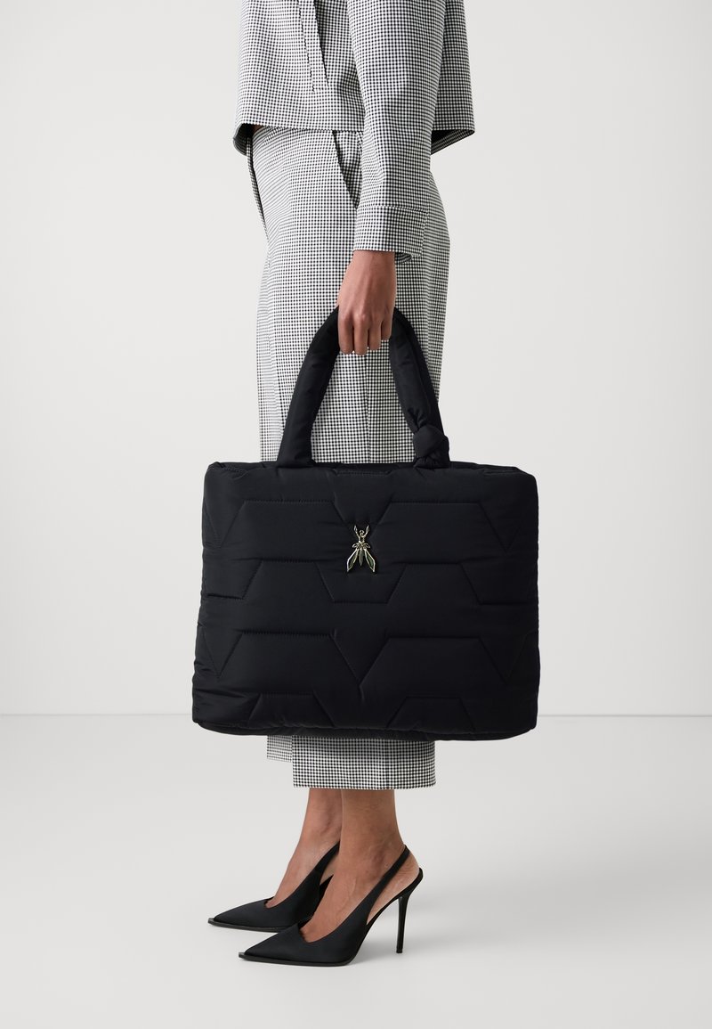 Personne portant un costume à carreaux et des talons hauts noirs tenant un grand sac fourre-tout noir matelassé avec un ornement en métal en forme de fourmi.