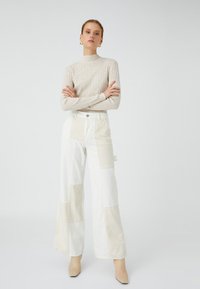 Beige ribattuto maglione a collo alto abbinato a pantaloni larghi in patchwork nei toni crema e beige chiaro. Il modello è in piedi con le braccia incrociate.