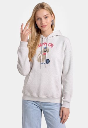 Ung kvinde med langt blondt hår iført en lys grå hoodie med en kat og garn grafik og blå jeans, smilende blidt.