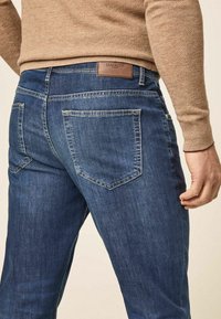 Hombre que lleva pantalones vaqueros azules con bolsillos traseros y una etiqueta de marca de cuero marrón, combinados con un suéter de punto beige.