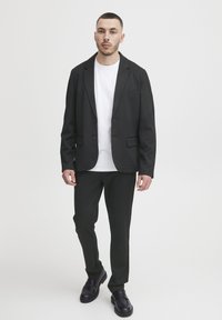 Schwarzer Blazer und Hose in Slim-Fit, mit glatter Textur, zwei vorderen Knöpfen und einem weißen T-Shirt darunter. Schwarze Schuhe ergänzen den Look.