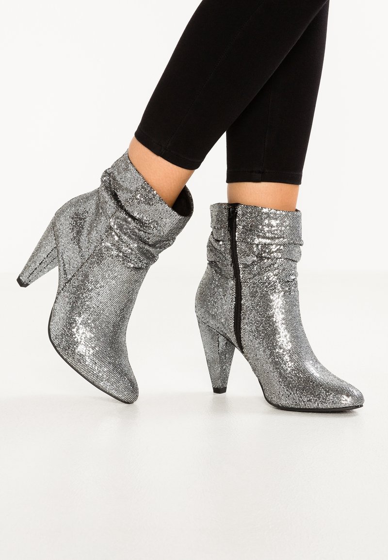 Bottes argentées pailletées jusqu'à la cheville, à bouts pointus, talons empilés, texture froissée, fermeture éclair latérale, portées avec un pantalon noir ajusté.