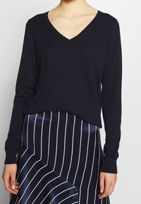 Granatowy sweter z dekoltem w serek, wykonany z miękkiego dzianego materiału, zestawiony z granatową spódnicą w białe prążki o lekko błyszczącej fakturze i asymetrycznym kroju.