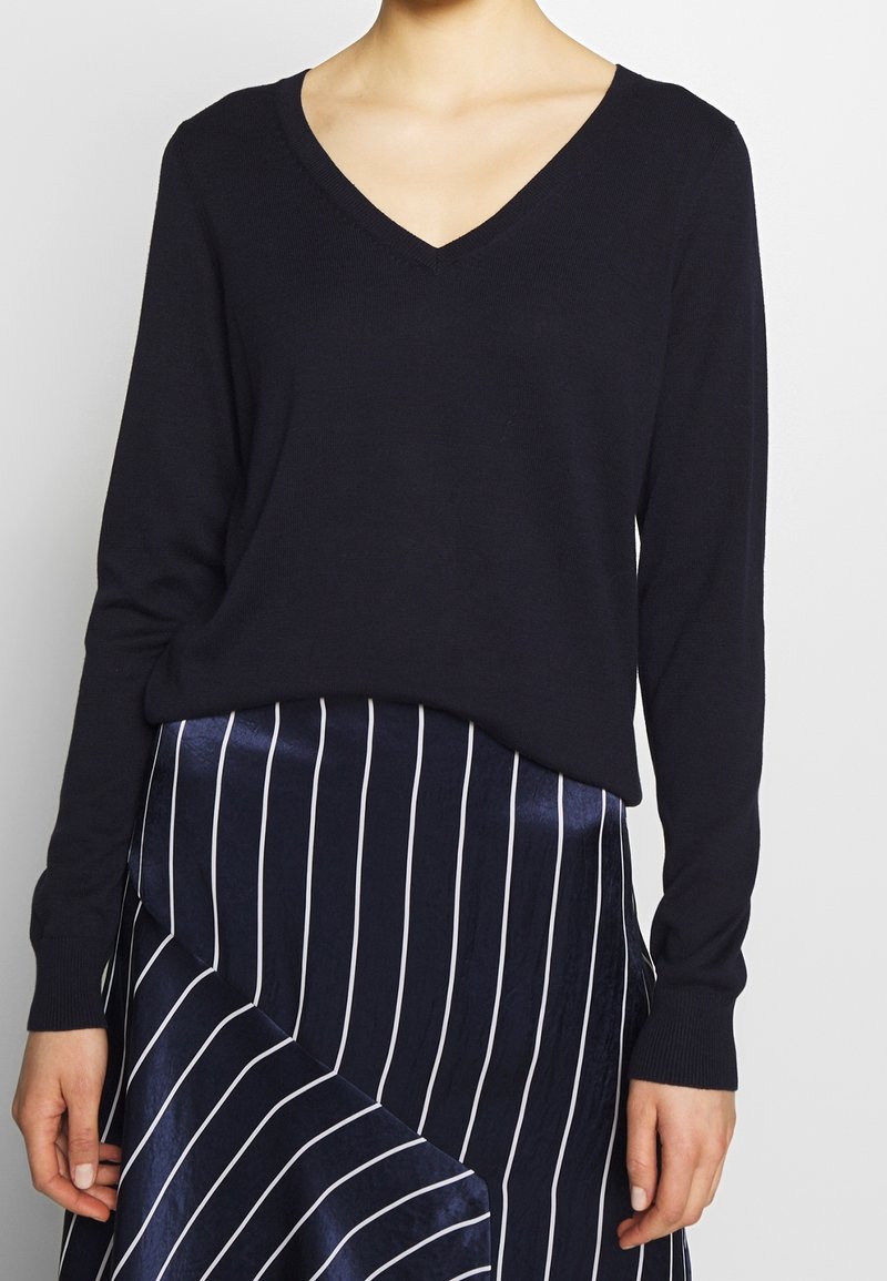 Granatowy sweter z dekoltem w serek, wykonany z miękkiego dzianego materiału, zestawiony z granatową spódnicą w białe prążki o lekko błyszczącej fakturze i asymetrycznym kroju.