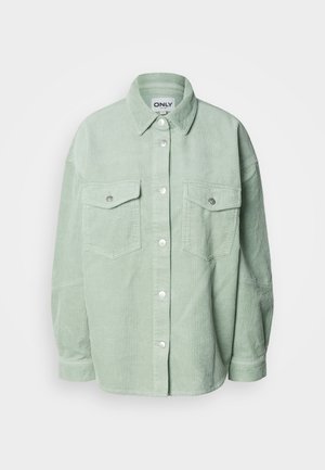 Gaiši zaļa kordurovas krekla ar divām krūšu kabatām ar aizvaru un garām piedurknēm, apkakle salocīta uz leju.