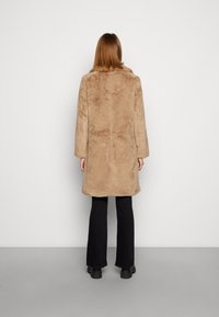 Beżowy płaszcz ze sztucznego futra z kołnierzem, długimi rękawami i lekko luźnym krojem, zestawiony z czarnymi rozkloszowanymi spodniami i czarnymi botkami.