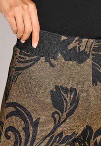 Een hand plaatst een vinger in de zak van een broek met een patroon van zwarte en gouden bloemmotieven en een zwarte tailleband.