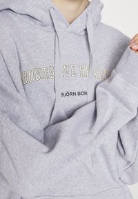 Sweat à capuche gris en tissu doux. Présente le texte "PROGRESSIVE HEALTH" en crème et le logo "BJÖRN BORG". Affiche une coupe décontractée.