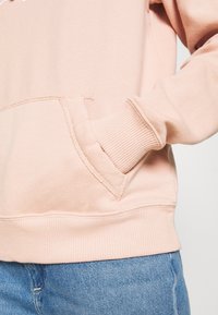 Jasnoróżowa bluza z kapturem z przednią kieszenią, ściągaczami na mankietach i u dołu. Materiał wydaje się miękki o gładkiej fakturze, odpowiedni do codziennego noszenia.