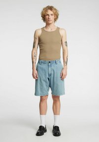 Brun topp, lyseblå denimshorts med oppbrettede kanter, hvite sokker, svarte loafers, tatoveringer synlige på armene, står foran en nøytral bakgrunn.