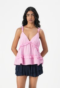 Rosa Baumwoll-Hemd ohne Ärmel mit Rüschen-Trägern, einem V-Ausschnitt und einem geschichteten Rüschen-Saum, kombiniert mit navy-blauen, plissierten Shorts.