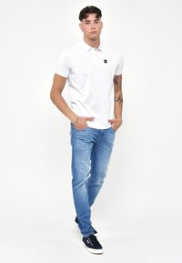 Polo bianco con un piccolo logo, abbinato a jeans azzurri e scarpe da ginnastica blu scuro. Il modello è in piedi, con le mani nelle tasche, su uno sfondo semplice.