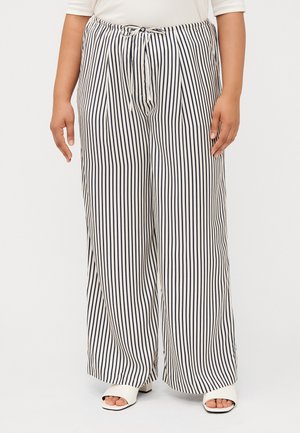 Une personne portant un pantalon large à rayures blanc et marine se tient debout, les mains détendues le long du corps, complétée par des chaussures blanches à bout ouvert.