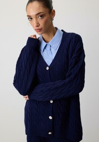 Marinebl�å strikket cardigan med kabelmønster, V-hals og lange ærmer. Har sølvknapper, struktureret stof og en afslappet pasform.