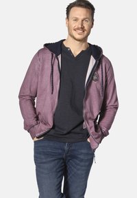 Purple zip-up hoodie with a navy hood, featuring a front emblem, paired with a black v-neck shirt and blue jeans. Smooth fabric texture.