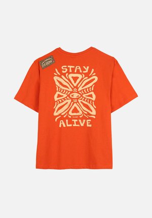 T-shirt en coton orange à manches courtes, présentant un grand motif graphique crème et le texte "STAY ALIVE" au dos. Étiquette sur l'épaule.