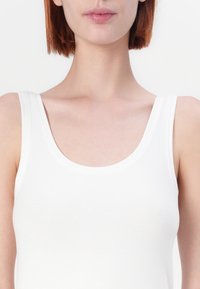 Balts bez piedurknēm tank tops ar apaļu kakla izgriezumu, izgatavots no gludas kokvilnas auduma. Raksturo tīrs, minimāls dizains un nav redzamu rakstu.