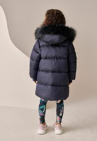 Kék puffertákó bunda, szőrmebélésű kapucnival, steppelt mintázattal. Színes, mintás leggings párosítva és fehér tornacipő.