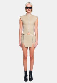 Beige Crop-Top mit kurzen Ärmeln und Frontreißverschluss, kombiniert mit einem passenden Minirock mit seitlichen Schlitzen und kontrastierenden Reißverschlüssen. Schwarze Ankle-Boots.