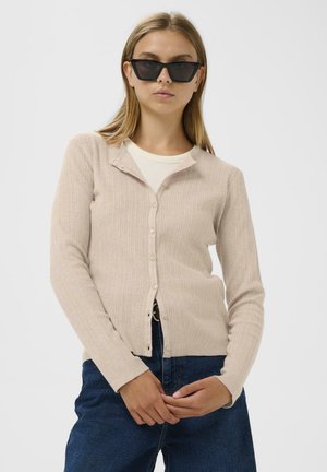 Beige bordás kardigán gombos elejével, slim fit szabással és hosszú ujjal, sötét farmernadrággal és fekete napszemüveggel párosítva.