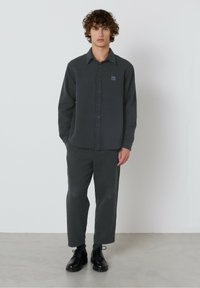 Mladý muž s kučeravými vlasmi, oblečený v tmavosivej košeli na gombíky a zodpovedajúcich nohaviciach, stojí na svetlosivej podlahe proti bielej stene.