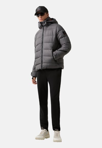Grå polstret jakke med hætte, lynlåslukning og sorte accenter. Båret med sorte bukser og lyse, kraftige sneakers.